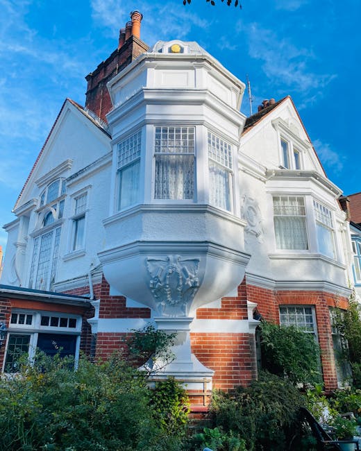 A street scene showing a white, historic period house with ornate architectural details, including a rounded bay window, decorative cornices, and a small balcony with black wrought iron railings. In f