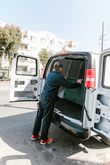 Inside the open rear doors of a moving van, two young women are sitting on the edge of the vehicle's floor, smiling and engaging in conversation. One woman, with curly hair and casual clothing, is ges