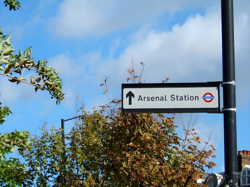 Photograph of a rectangular directional sign mounted on a black metal pole, pointing straight ahead towards Arsenal Station. The sign displays a white background with black text and includes the Londo