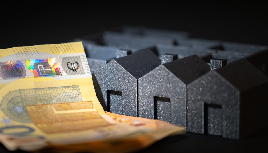 Close-up image showing a yellow 50-pound banknote partially draped over small, dark grey foam house-shaped models, which are arranged in a row on a black surface. The foam houses have textured surface