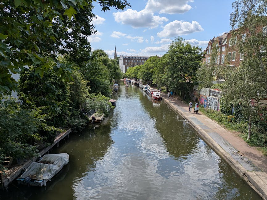 A scenic view of the Upper Street Islington area showing a narrow waterway with calm, reflective water, flanked by lush green trees on both sides. On the right, there is a paved sidewalk with pedestrians walking, alongside residential buildings with brick facades. Parked cars line the street, and a graffiti-covered wall is visible among the foliage. The sky is partly cloudy with patches of blue, indicating a bright day. In the distance, the church spire rises above the treetops, contributing to the urban landscape. This setting illustrates the typical environment where home relocation and furniture transport services by Man With a Van Islington might operate, especially when navigating narrow access properties or working within urban landscapes during packing and moving processes.