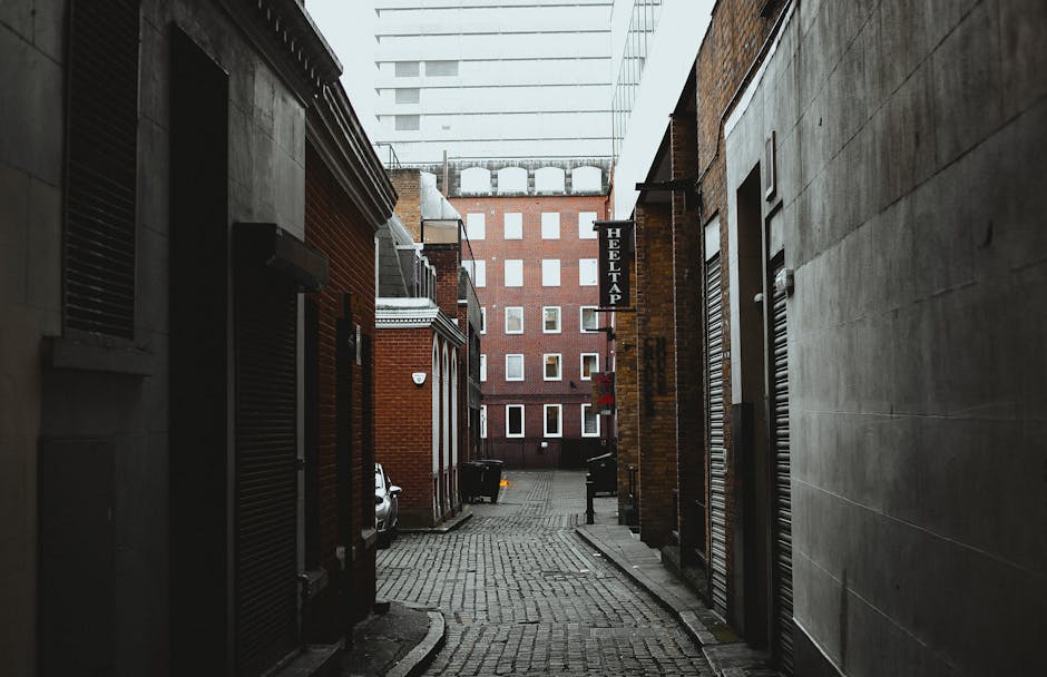 A narrow cobblestone alleyway in Islington, flanked by brick and concrete buildings, with a small white car parked partially visible on the left side. Several doors with metal roller shutters are closed along the alley; one has a sign for a hotel tap. The alley is under an overcast sky, with modern multi-storey office or apartment buildings visible at the end of the passage. This scene captures the typical urban environment where house removals and furniture transport may be coordinated by a professional service like Man With a Van Islington, dealing with logistics and tight access for home relocation or moving services.