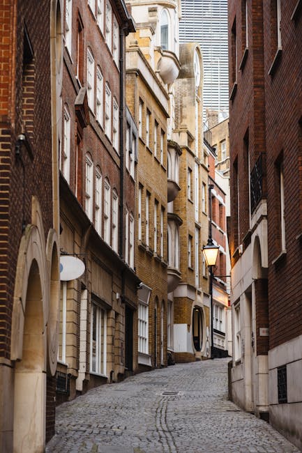 A scenic view of the Upper Street Islington area showing a narrow waterway with calm, reflective water, flanked by lush green trees on both sides. On the right, there is a paved sidewalk with pedestrians walking, alongside residential buildings with brick facades. Parked cars line the street, and a graffiti-covered wall is visible among the foliage. The sky is partly cloudy with patches of blue, indicating a bright day. In the distance, the church spire rises above the treetops, contributing to the urban landscape. This setting illustrates the typical environment where home relocation and furniture transport services by Man With a Van Islington might operate, especially when navigating narrow access properties or working within urban landscapes during packing and moving processes.